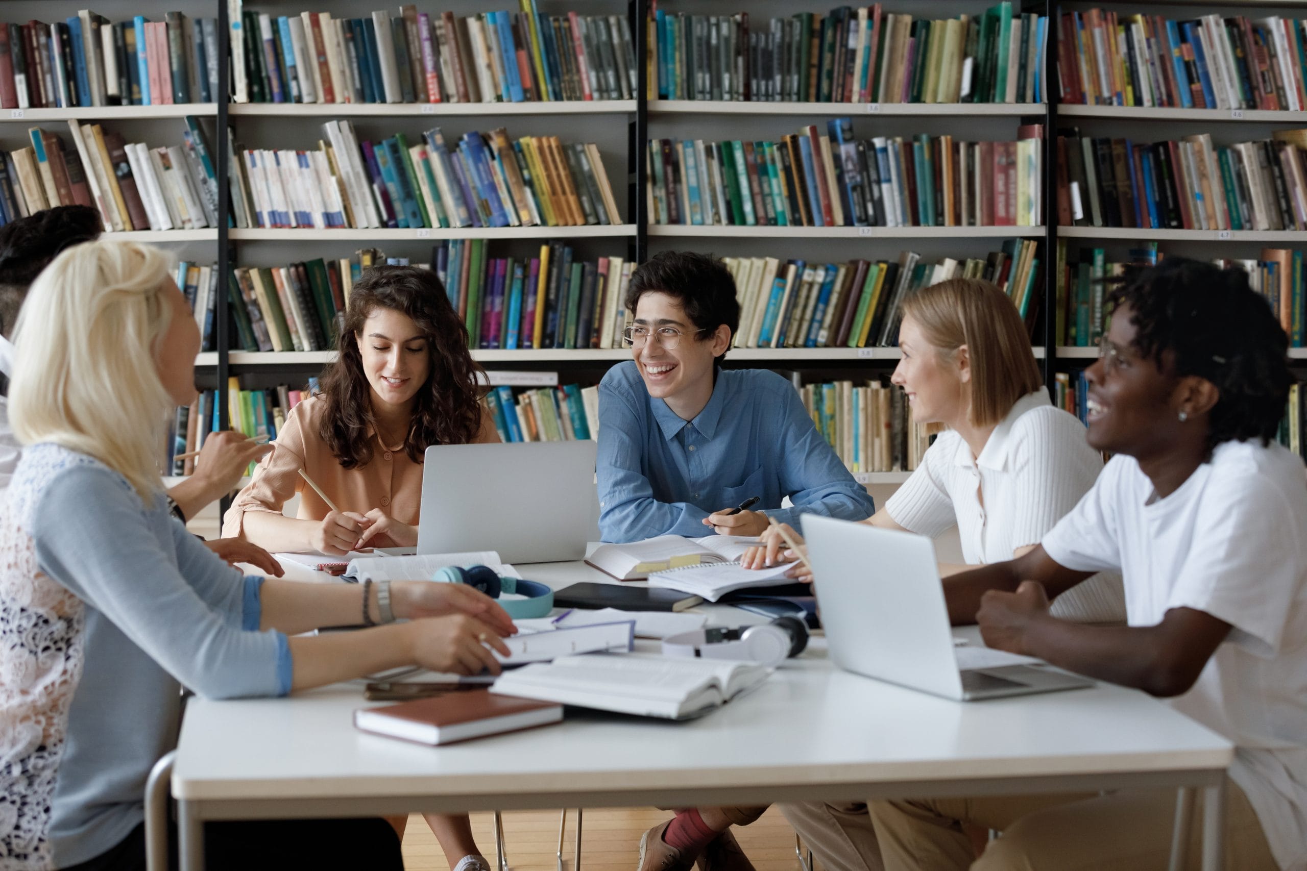 A diverse group of college students sit at a study table in a library, with shelves of books in the background