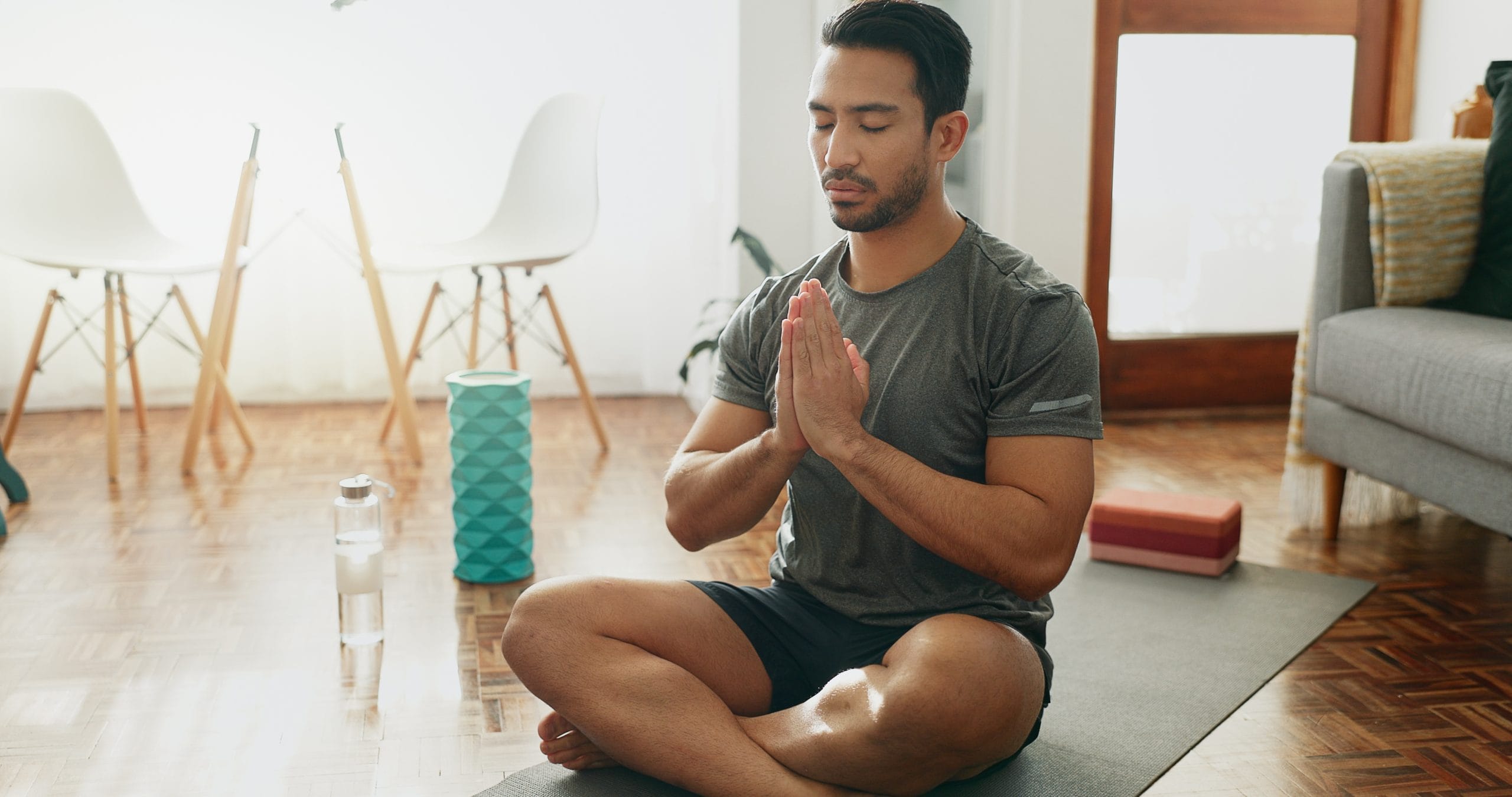 A man with dark hair and a beard sits cross-legged on a yoga mat with his hands in prayer position and his eyes closed