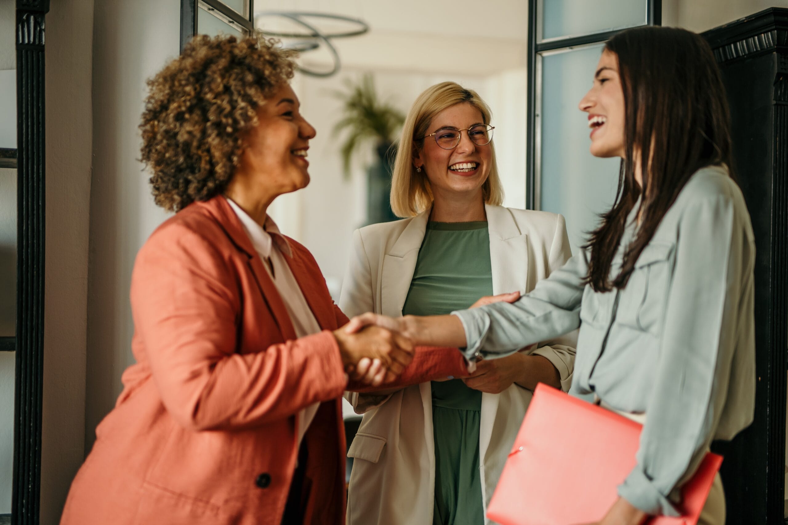 Three women in business attire smile at one another, with the two in the foreground shaking hands