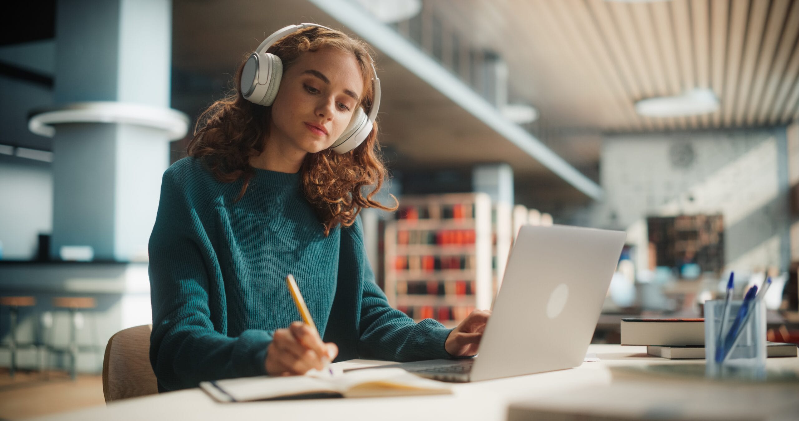 A young woman with curly hair sits at a table in a library while taking notes on a laptop and wearing white headphones
