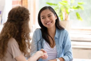 A young woman in a blue chambray shirt smiles and shakes hands with another woman