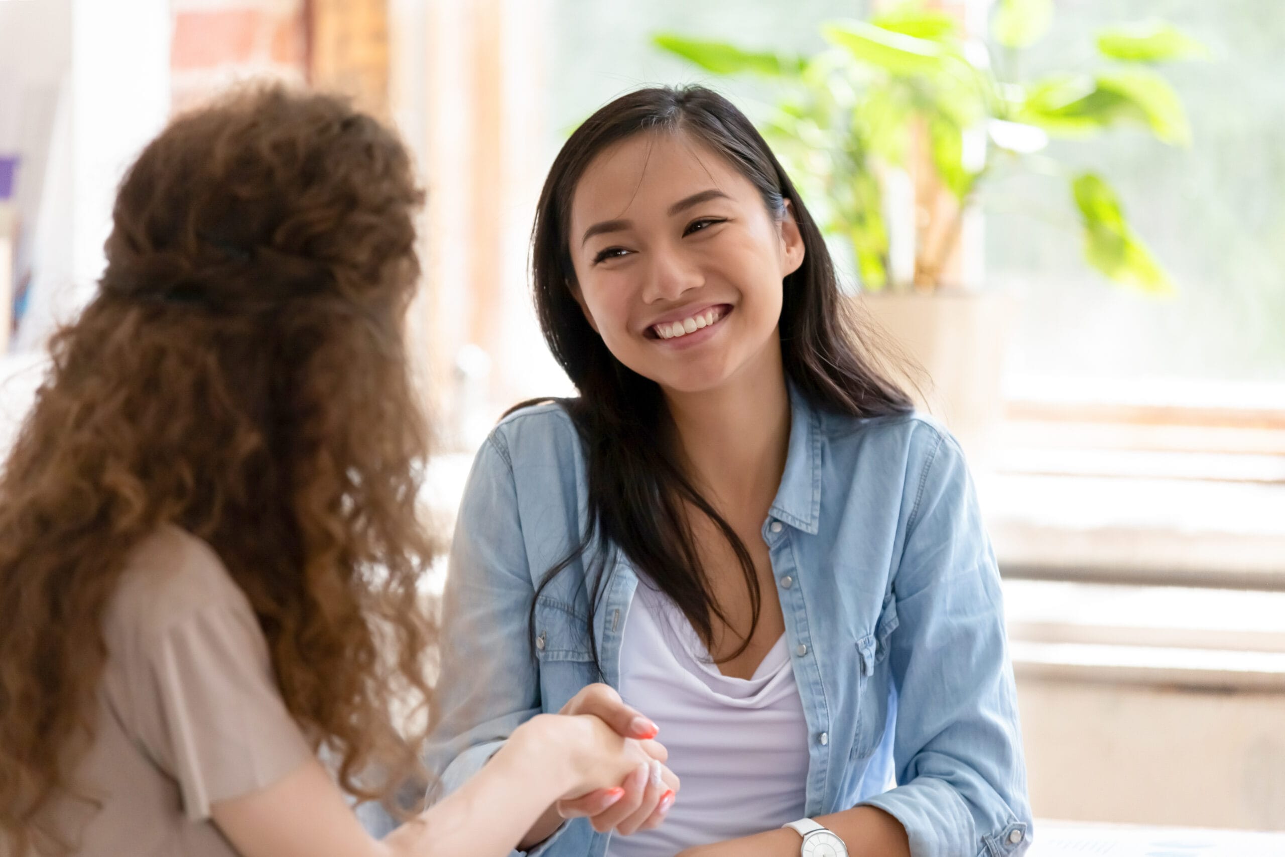 A young woman in a blue chambray shirt smiles and shakes hands with another woman